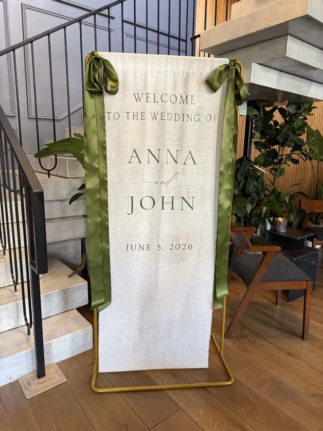 Printed fabric welcome banner reading Welcome to the wedding of Anna and John, hung on a gold stand with olive green ribbon ties