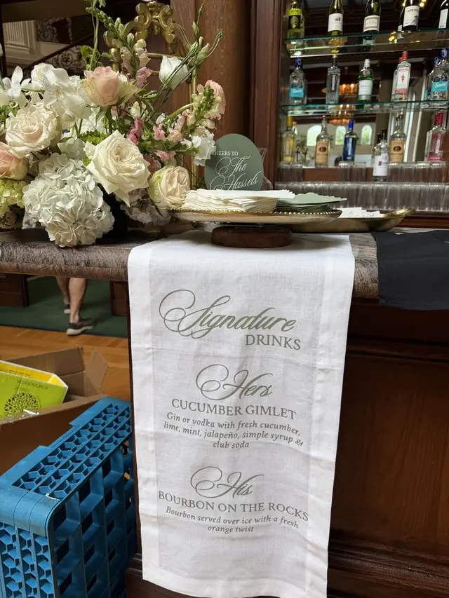 Printed fabric signature drinks runner draped over a wedding bar beside a white floral arrangement