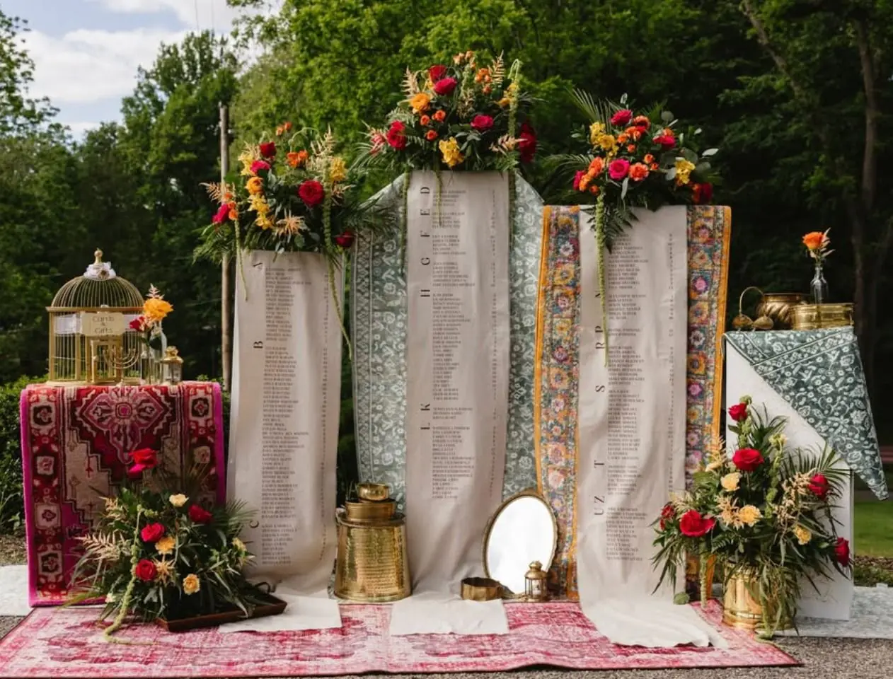 A large outdoor wedding seating chart display made of printed fabric panels, styled with vintage rugs, brass lanterns, and bright florals
