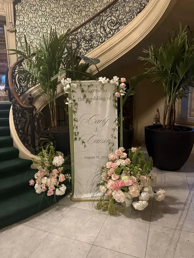 Fabric welcome banner for Carly and Carson displayed on a gold easel beside blush florals at the foot of a grand staircase